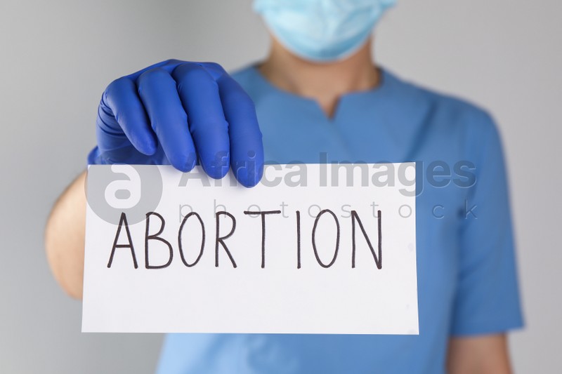 Doctor holding paper card with word ABORTION on light grey background, closeup