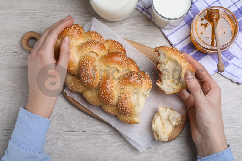Photo of Woman breaking freshly baked braided bread with sesame seeds at wooden table, top view. Traditional Shabbat challah