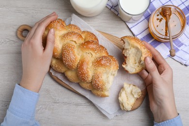 Woman breaking freshly baked braided bread with sesame seeds at wooden table, top view. Traditional Shabbat challah