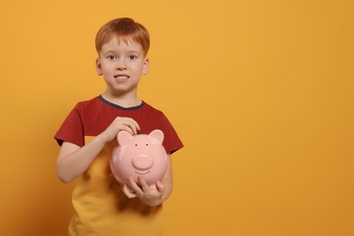 Cute little boy with ceramic piggy bank on orange background, space for text