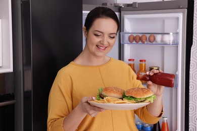 Photo of Happy overweight woman with ketchup and burgers near fridge in kitchen
