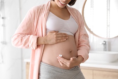 Photo of Young pregnant woman applying cosmetic product on belly in bathroom, closeup