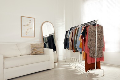 Rack with stylish clothes near sofa and mirror indoors. Fast fashion
