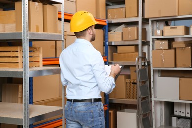 Young man with clipboard near rack of cardboard boxes at warehouse, back view