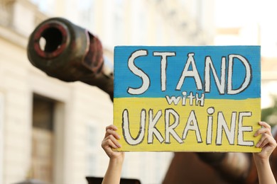 Woman holding poster in colors of national flag with words Stand with Ukraine near broken tank on city street, closeup