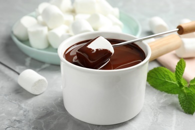 Dipping marshmallow into fondue pot with dark chocolate on marble table, closeup