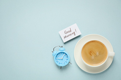 Delicious coffee, alarm clock and card with GOOD MORNING wish on light background, flat lay. Space for text