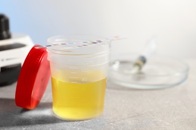 Container with urine sample for analysis and test strips on grey table in laboratory, closeup. Space for text