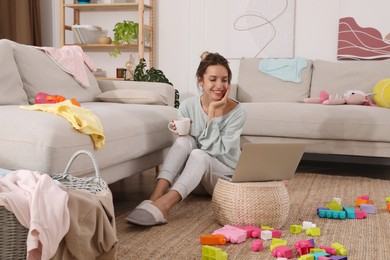 Young mother drinking tea and working with laptop on floor in messy room