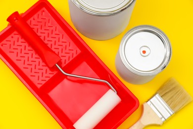 Photo of Cans of orange paint, brush, roller and container on yellow background, above view