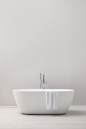 Modern ceramic bathtub with towel near light wall indoors