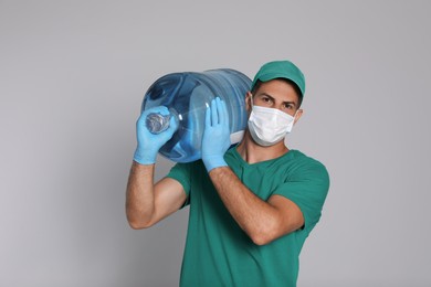 Courier in medical mask holding bottle for water cooler on light grey background. Delivery during coronavirus quarantine