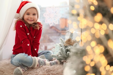 Little girl in Santa hat near small Christmas tree decorated with snowflakes at home