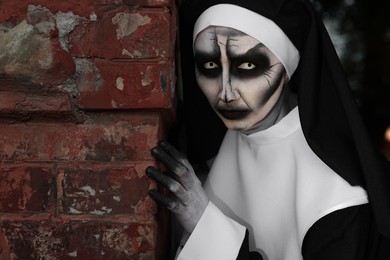 Scary devilish nun hiding behind red brick wall outdoors. Halloween party look