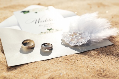 Envelope with wedding invitation, brooch and gold rings on sandy beach, closeup