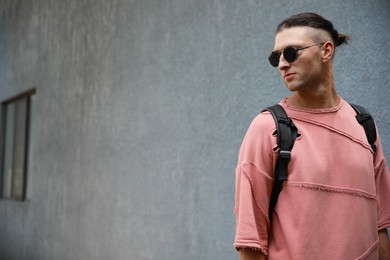 Handsome young man in stylish sunglasses with backpack near grey wall outdoors, space for text