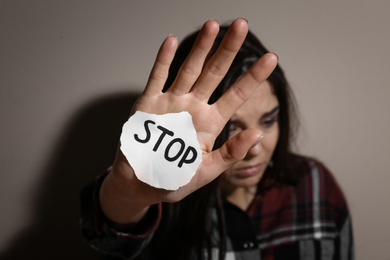 Abused young woman with sign STOP near beige wall, focus on hand. Domestic violence concept