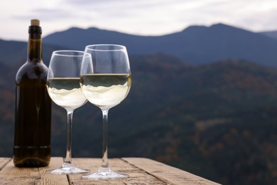 Photo of Glasses and bottle of tasty wine on wooden table against mountain landscape. Space for text