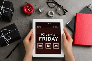 Woman with tablet surrounded by gifts and accessories at grey table, top view. Black Friday sale