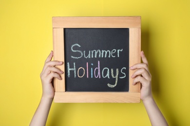 Photo of Schoolgirl holding blackboard with text SUMMER HOLIDAYS on yellow background, closeup