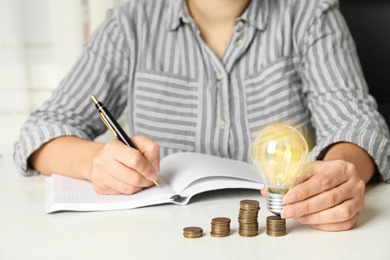 Woman with light bulb, notebook and coins at white table, closeup. Power saving