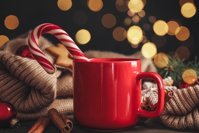 Cup of tasty cocoa with Christmas candy cane on wooden table against blurred festive lights