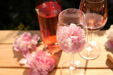 Bottle and glasses of rose wine near beautiful peonies on wooden table outdoors