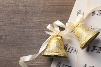 Photo of Golden shiny bells with bows and music sheets on wooden table, flat lay. Space for text. Christmas decoration