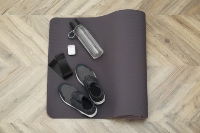 Exercise mat, bottle of water, wireless earphones, fitness elastic band and shoes on wooden floor, top view