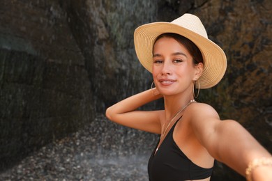 Beautiful young woman in stylish bikini taking selfie near waterfall outdoors, space for text