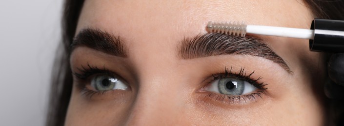 Beautician fixing woman's eyebrows with gel after tinting on light grey background, closeup