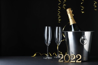 Happy New Year 2022! Bottle of sparkling wine in bucket and glasses on table against black background, space for text