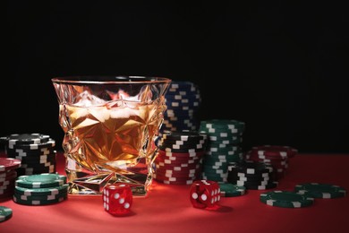 Casino chips, dice and glass of whiskey on red table against black background, space for text