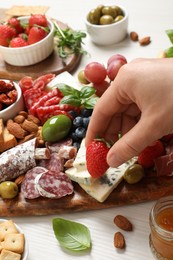 Woman taking strawberry from board with different appetizers at white wooden table, closeup