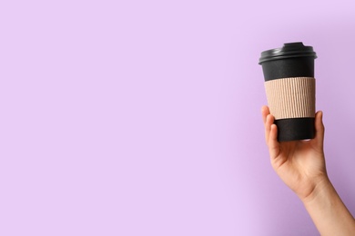 Woman holding takeaway paper coffee cup with cardboard sleeve on violet background, closeup. Space for text