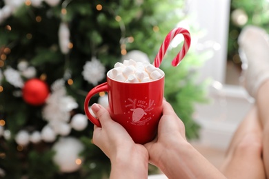 Woman with cup of delicious hot drink near Christmas tree indoors, closeup