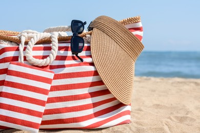 Stylish striped bag with visor cap and sunglasses on sandy beach near sea, closeup