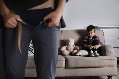 Man with unzipped pants standing near scared little boy on sofa indoors. Child in danger
