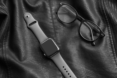 Stylish smart watch and glasses on black leather fabric, above view
