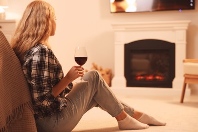 Photo of Beautiful young woman with glass of wine resting near fireplace at home