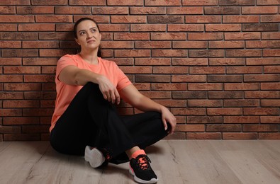 Overweight woman sitting near brick wall indoors, space for text