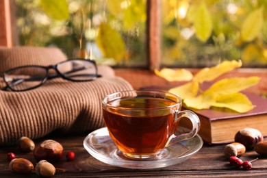 Photo of Cup of aromatic tea, acorns and soft sweater on wooden windowsill indoors. Autumn atmosphere