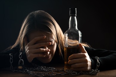 Alcohol addiction. Woman chained with bottle of whiskey at wooden table against black background