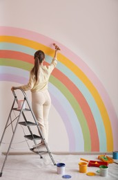 Young woman painting rainbow on white wall indoors, back view
