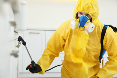 Pest control worker in protective suit spraying insecticide on furniture indoors