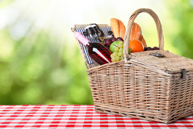 Wicker picnic basket with different products on checkered tablecloth outdoors, space for text