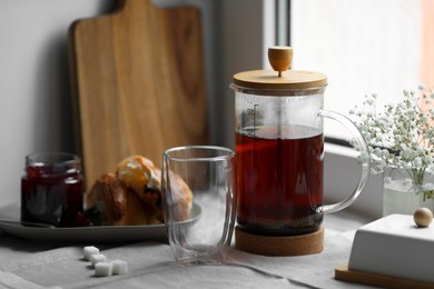 Teapot with freshly brewed tea, empty glass and sugar cubes on table near window