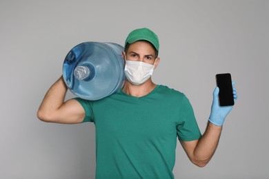 Courier in medical mask with bottle for water cooler showing mobile phone on light grey background. Delivery during coronavirus quarantine
