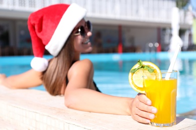 Young woman wearing Santa Claus hat with refreshing drink in swimming pool, focus on hand. Christmas vacation