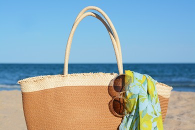 Photo of Straw bag with beach wrap and sunglasses on sandy seashore, closeup. Summer accessories